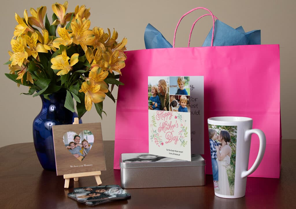 Flowers in vase, large gift bag, photo tile, tin, blank card and mug on table