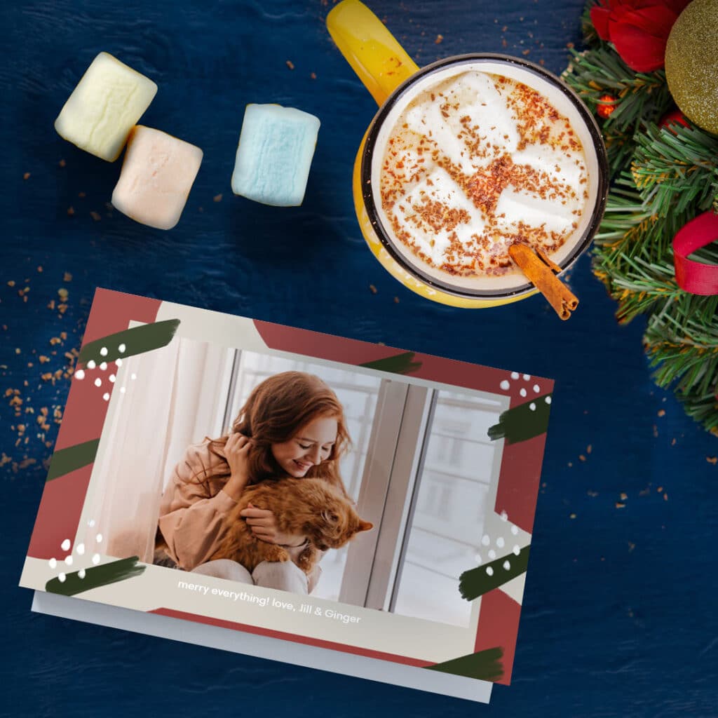 greeting card featuring wan cuddling with cat in front f laptop, surrounded by hot cocoa and marshmallows